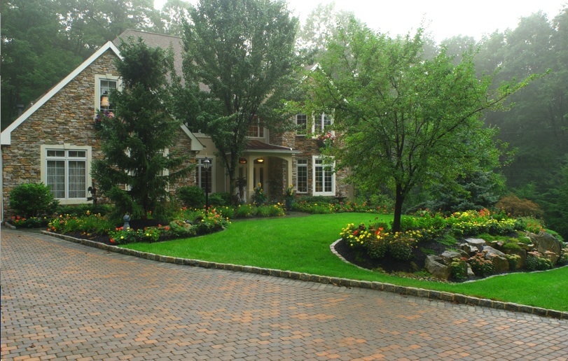 Picture of the front of a home, with flowers, shrubs and mainly trees as a part of the landscaping.